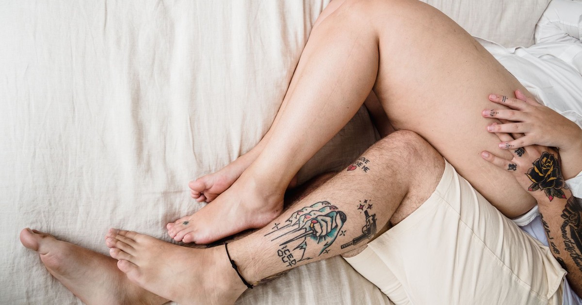 Close-up of a couple's tattooed legs intertwined on the bed in daylight.