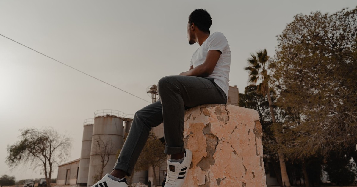 A man in casual wear sits on a stone structure outdoors, looking away.