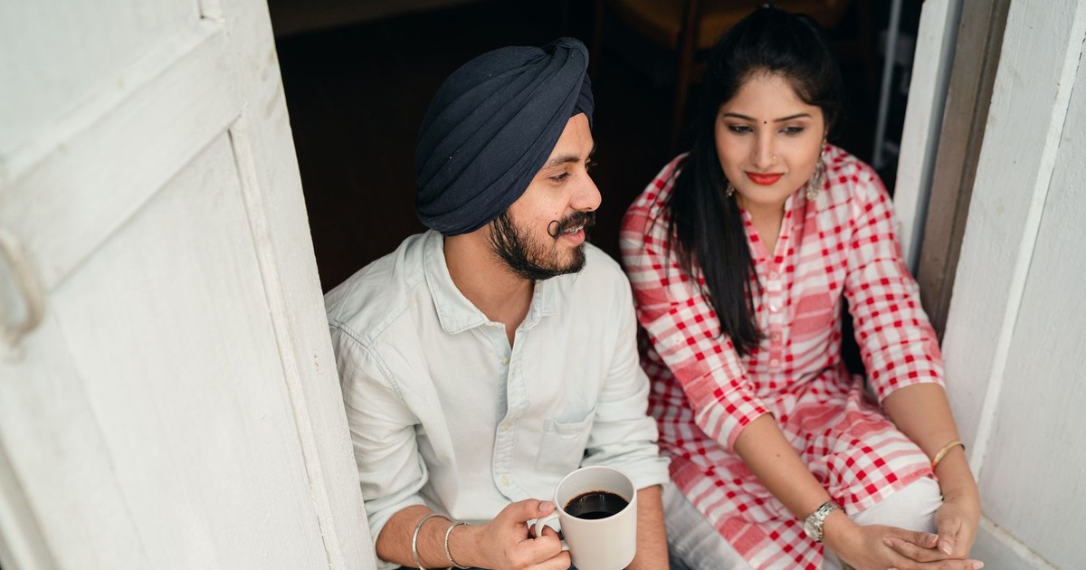 From above of good looking Indian wife listening to husband sharing news with cup of coffee in hand while both sitting at doorstep of house