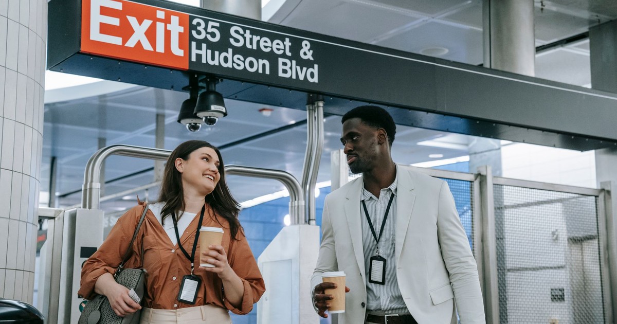 Man and woman exiting metro station in New York City, smiling and holding coffee cups.