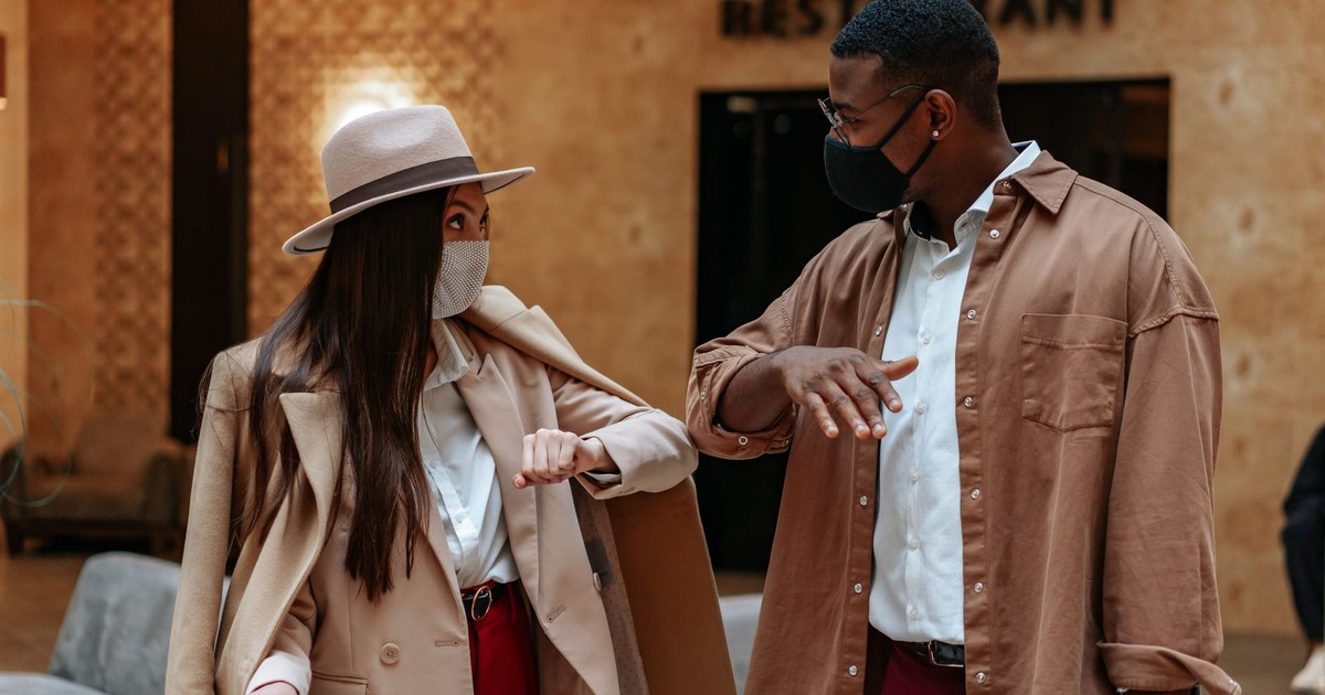 African American man and Hispanic woman greet each other with an elbow bump wearing face masks indoors.