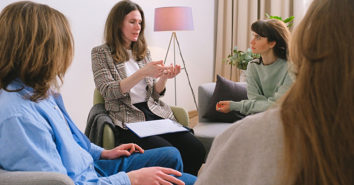 Young female therapist sitting on chair and discussing problem with patients during psychotherapy session in group
