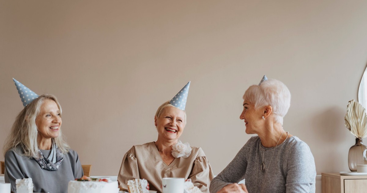 Three senior women enjoy a cozy indoor birthday celebration with party hats and smiles.