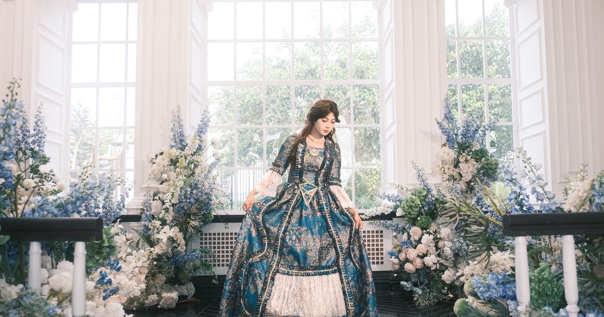 A woman in a blue Victorian dress stands amidst elegant floral decor indoors.