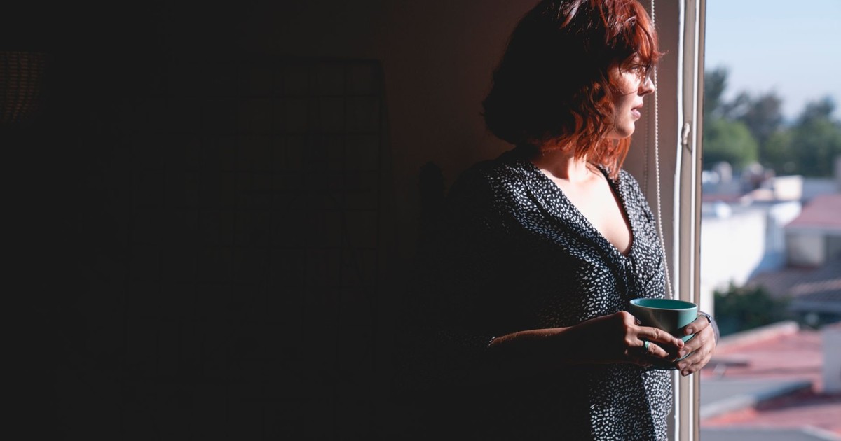 A woman stands by a window, holding a mug and lost in thoughtful contemplation.