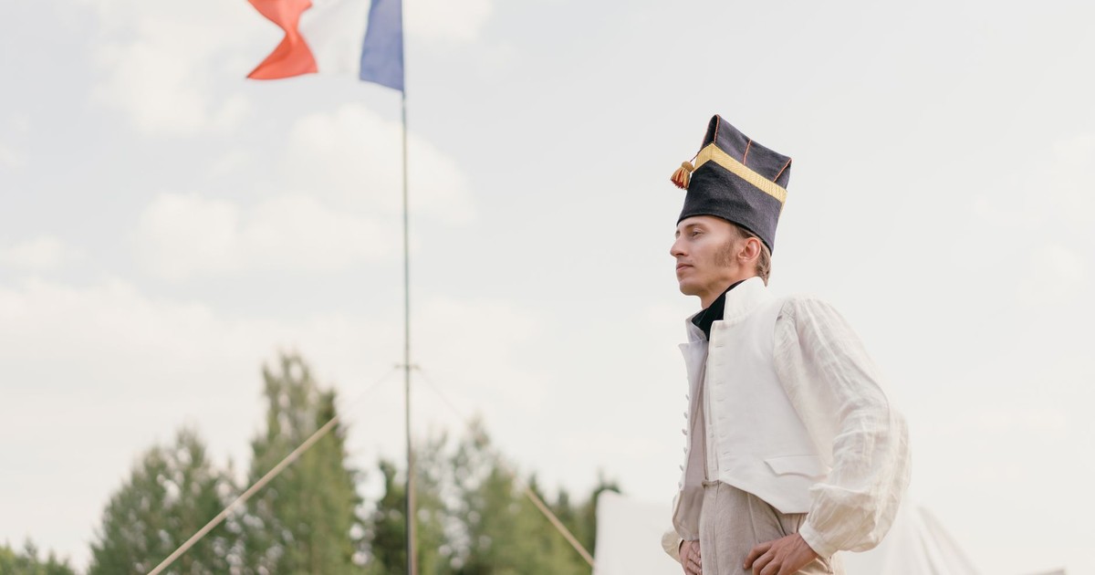 French soldier reenactor stands proud with tricolor flag in the background, symbolizing patriotism.