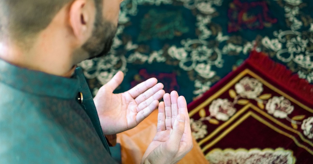 A man in traditional attire is praying on a beautifully decorated prayer mat indoors.