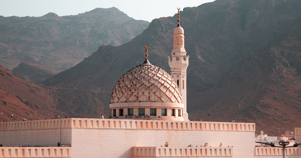 Scenic view of a mosque with domed roof and minaret set against a mountainous backdrop in Saudi Arabia.