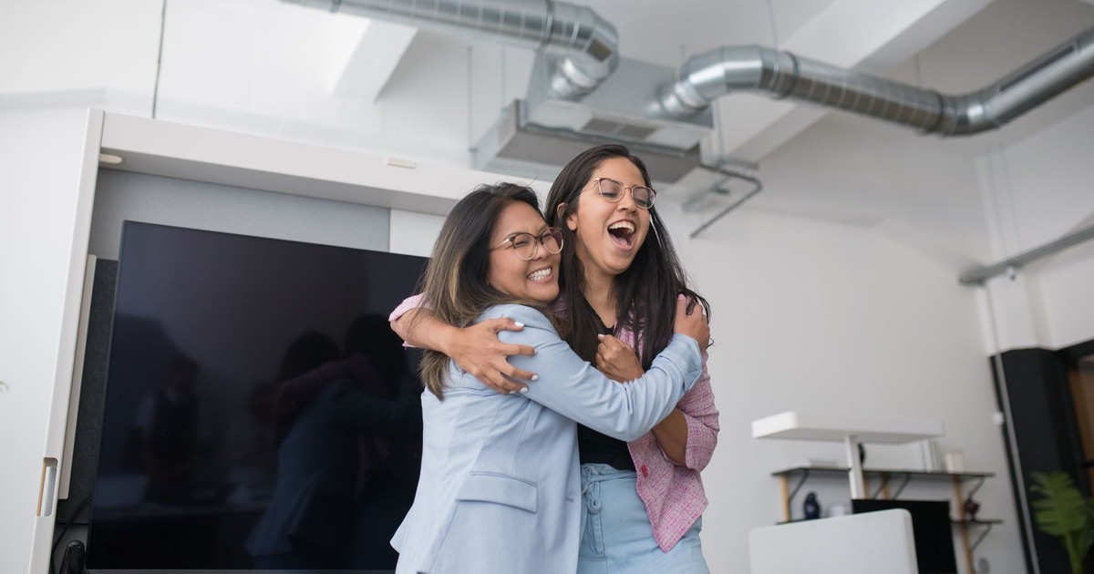 Two women confidently embrace in a bright modern office, expressing happiness and collegiality.