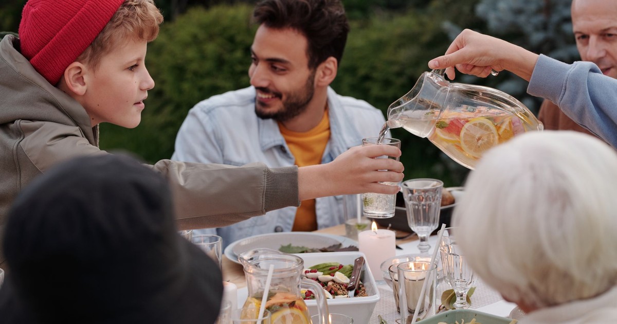 Faceless female pouring fruit beverage into glass of boy in warm cloth while cheerful multiethnic family having dinner outdoors