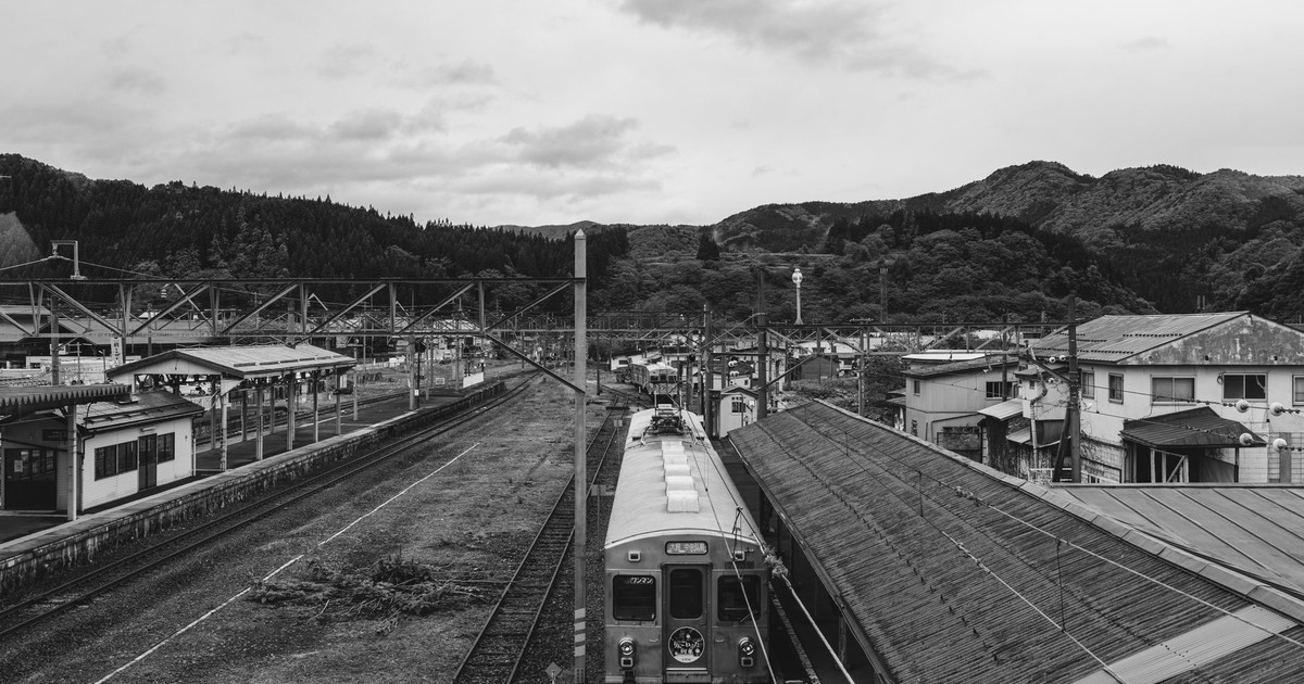 A vintage black and white photo of a rural train station with a scenic mountain backdrop.