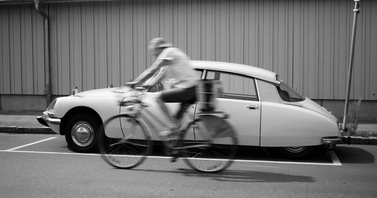 A vintage car parked on a city street with a cyclist in motion blur passing by.