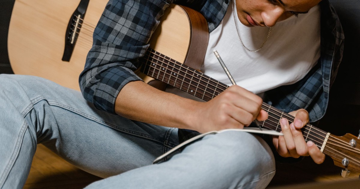 A young musician writes lyrics with a guitar by his side, deep in thought.