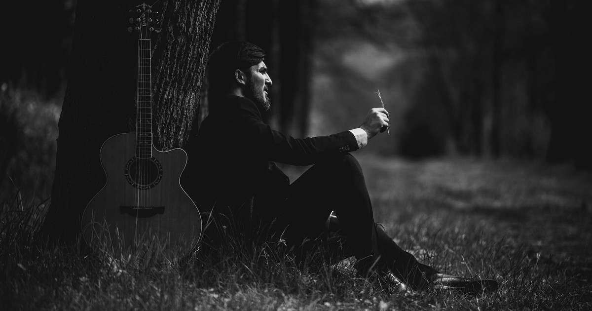Black and white image of man with guitar under tree. Contemplative atmosphere.
