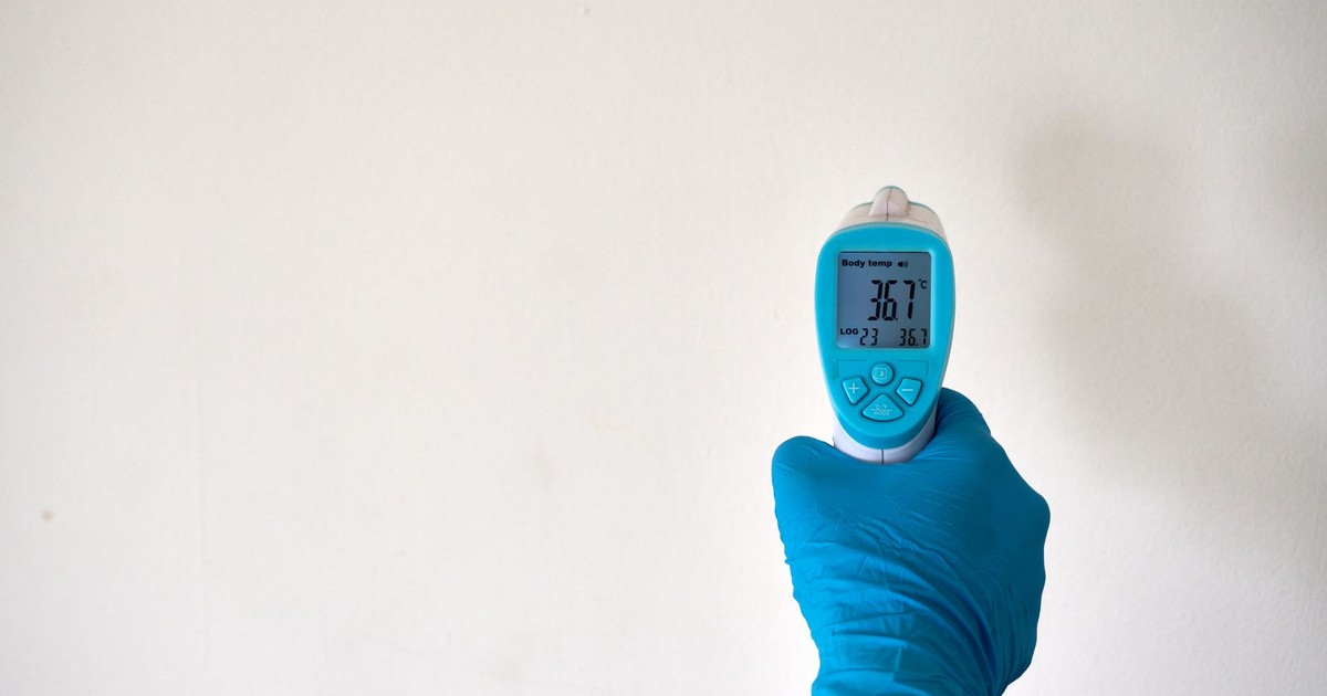 A hand in blue gloves holding an infrared thermometer against a plain background, signifying safety and health measures.