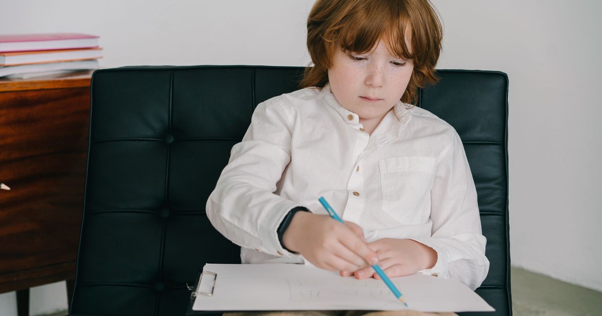 Child with red hair concentrates on drawing with colored pencils while sitting on a black chair.