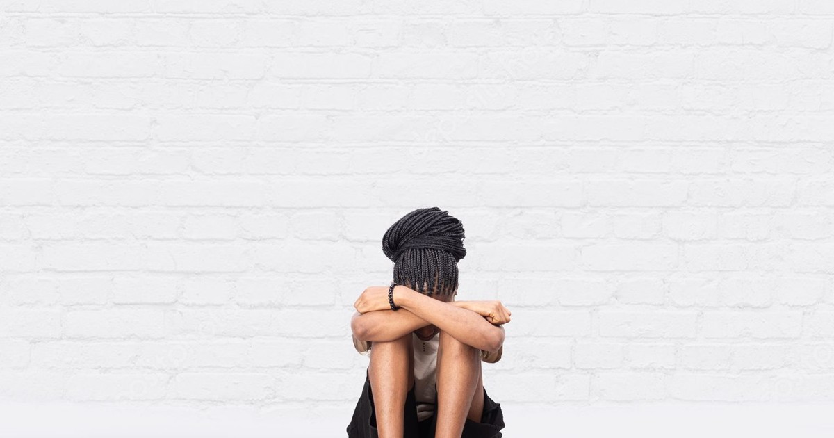 A woman with braided hair sitting with arms crossed, expressing emotion against a white brick wall.