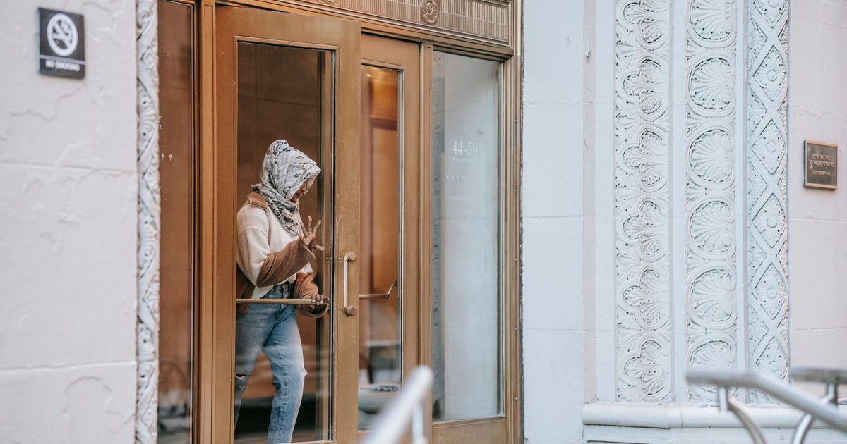 Side view of unrecognizable stylish Muslim lady in casual outfit and traditional headscarf leaving building with wooden doors and ornamental walls