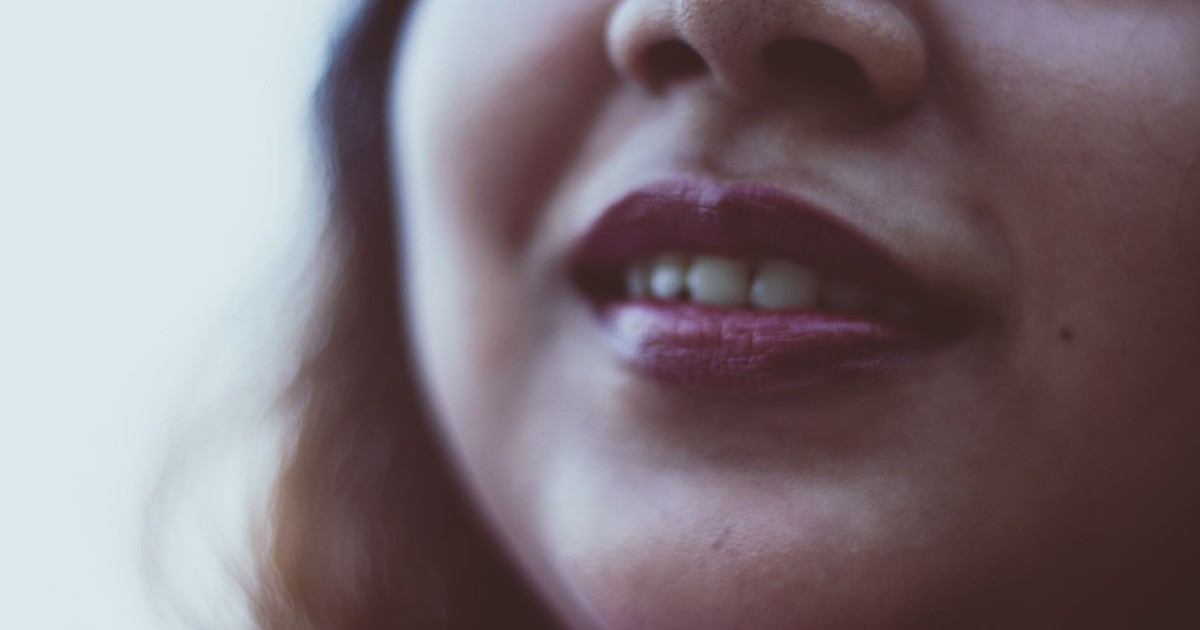 Closeup face of of unrecognizable cheerful female with dark loose hair with bright lipstick and toothy smile on blurred background in daylight