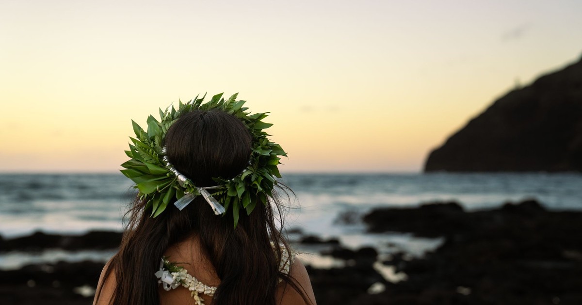 A woman wearing a lei and floral decoration gazes at the ocean during sunset, capturing a tranquil beach moment.