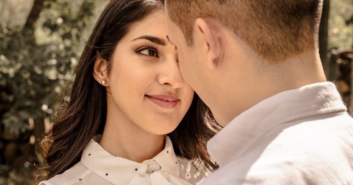 A young couple stands face to face, sharing a loving moment under a tree in daylight.