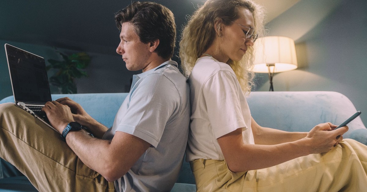 A young couple sits back-to-back, multitasking on digital devices in a cozy living room.