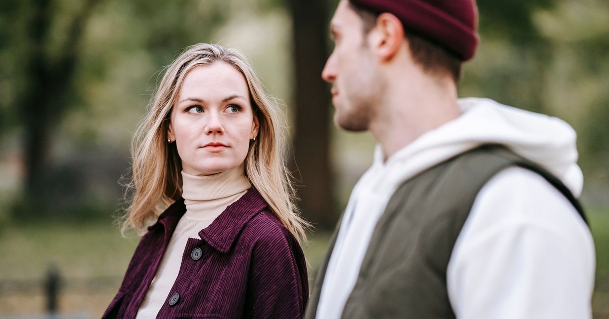 Calm woman in casual warm clothes with long hair looking at boyfriend with beard in hat while walking in park