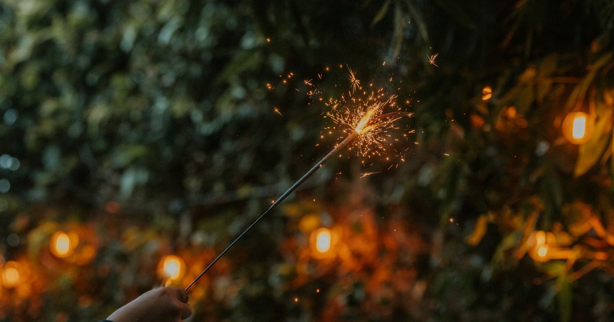 A hand holding a sparker illuminating the night, surrounded by festive outdoor lights.