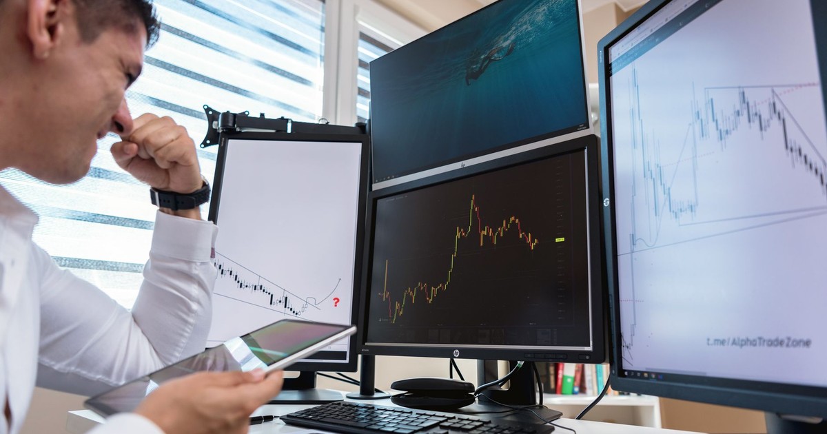 Trader in white shirt analyzing stock charts on multiple monitors during daytime in an office setting.
