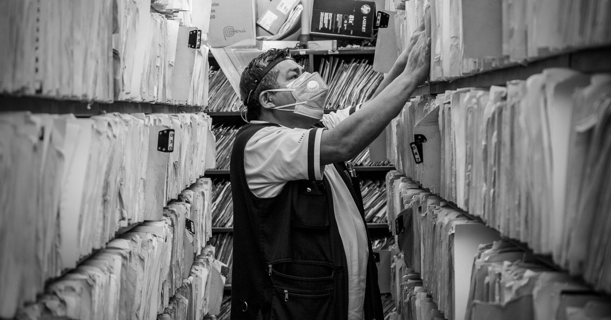 Elderly man sorts through files in an office aisle wearing a face mask.