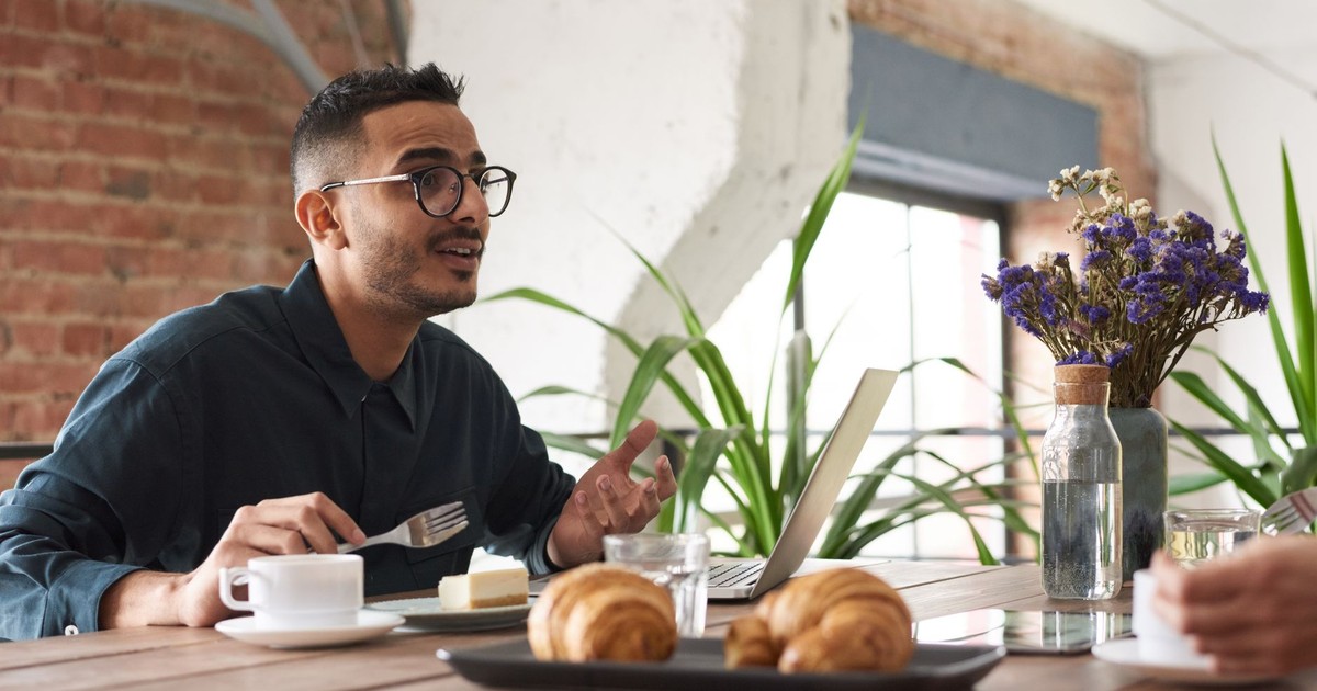 A man in glasses engaging in conversation over brunch with croissants and coffee.