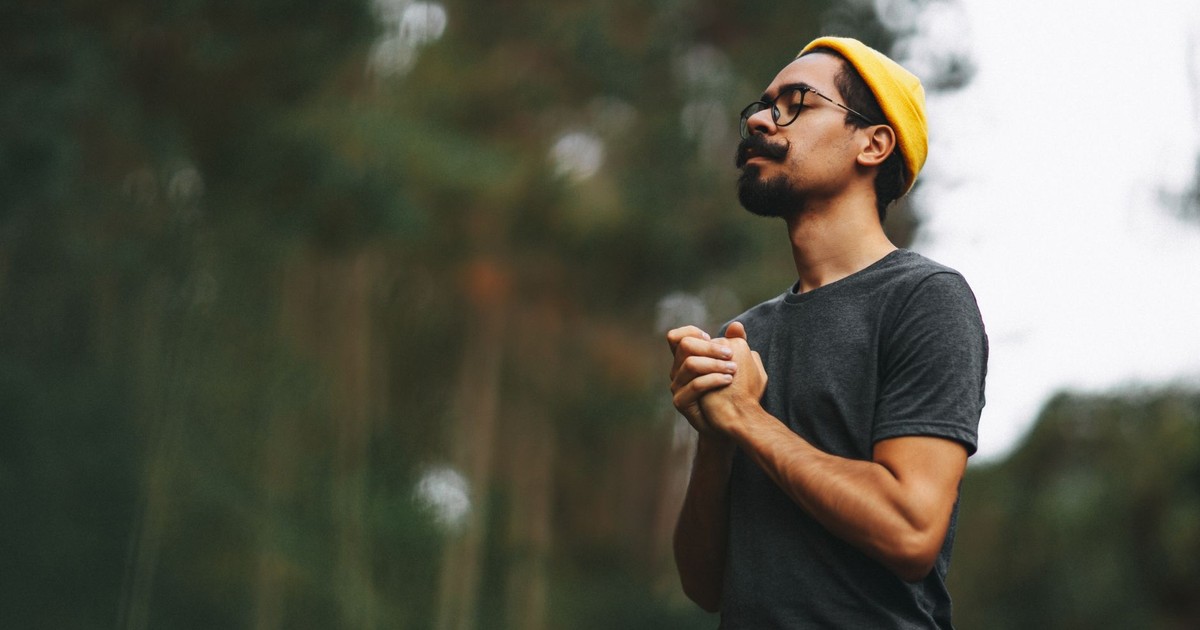 Portrait of a man with glasses and yellow beanie clasping hands outdoors.