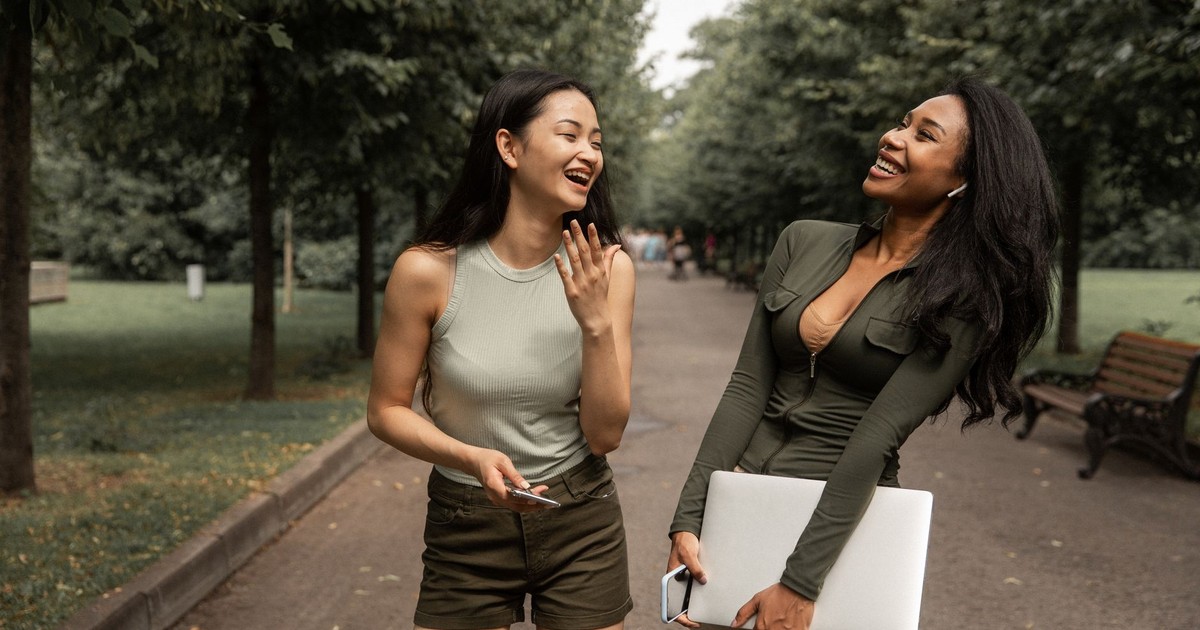 Happy young diverse female friends with smartphone and laptop in hands in casual clothes standing on path amidst lush trees in park and laughing while spending free time together