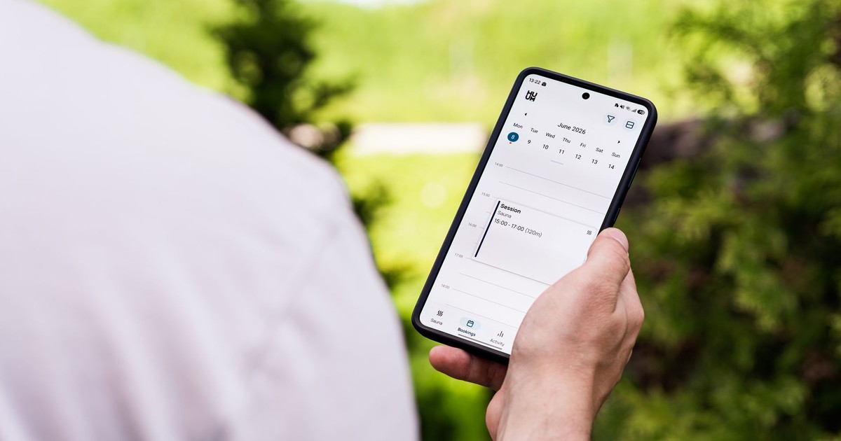 A person using a sauna booking app on a smartphone outdoors, surrounded by greenery.