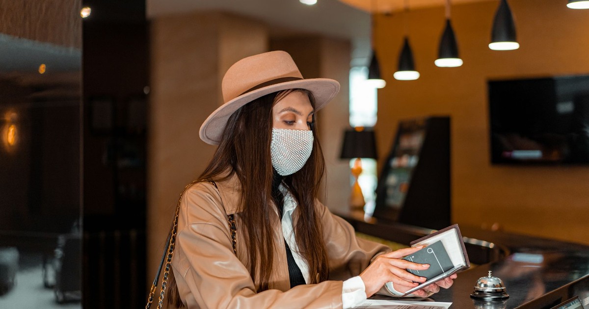 Woman wearing mask and hat checking in at hotel front desk, symbolizing travel and safety.