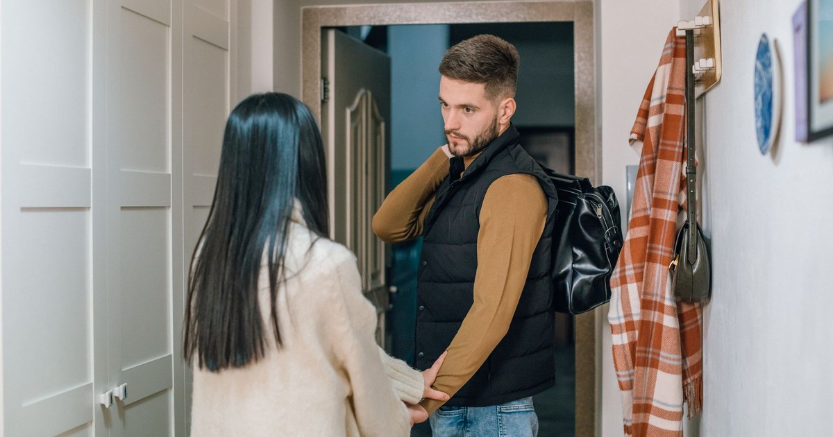 A couple holds hands in a hallway as a man prepares to leave with a backpack.