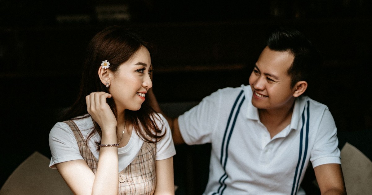 Cheerful shy Asian female in casual clothes with flower behind ear spending time with boyfriend together in city cafe