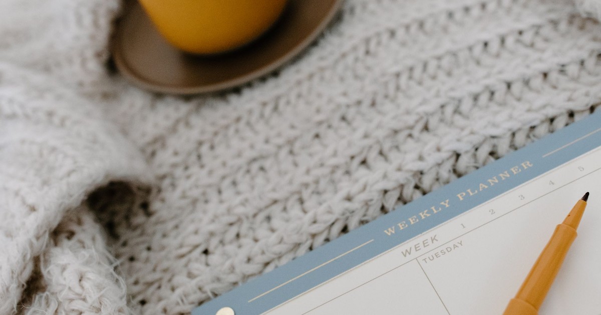 A cozy flat lay setup with a weekly planner, pen, and warm drink on a knitted blanket.