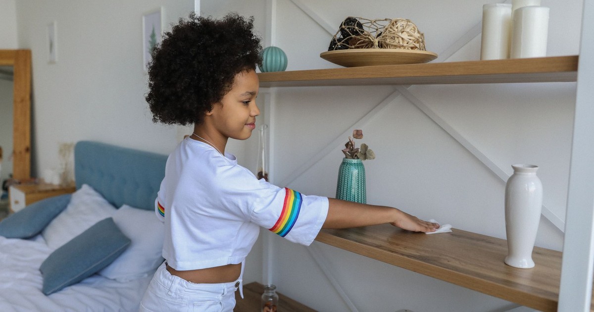 Charming African American girl wiping dust from wooden shelf of rack at home