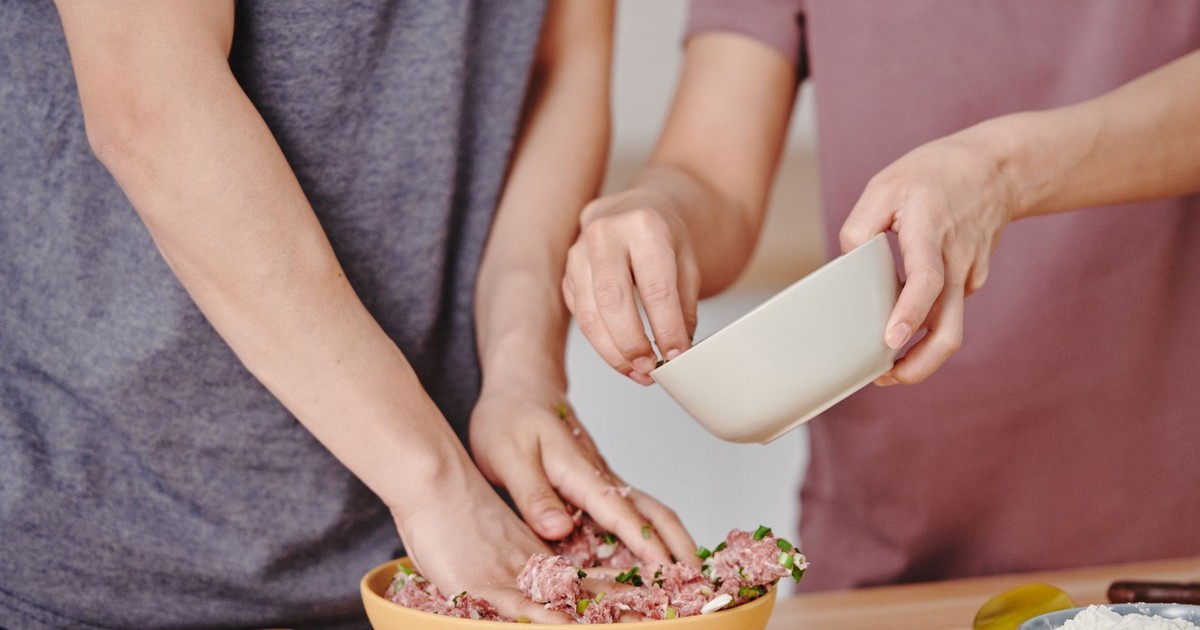 Two adults preparing a meat dish with fresh ingredients in a home kitchen.