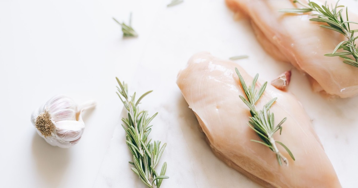 Raw chicken breasts garnished with rosemary and garlic on a white surface.