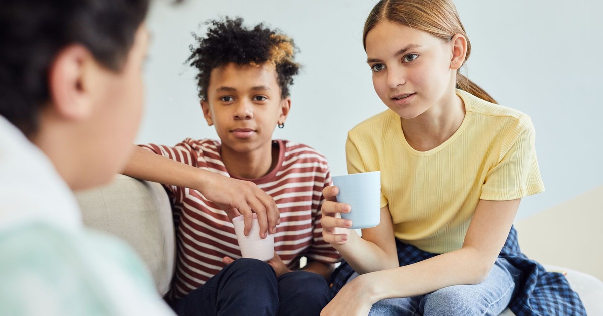 Group of positive diverse children sitting on sofa and holding cups of drink while spending time together against white wall