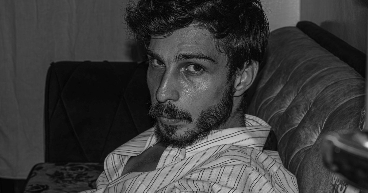 Moody black and white portrait of a bearded man in a striped shirt relaxing on a couch indoors.