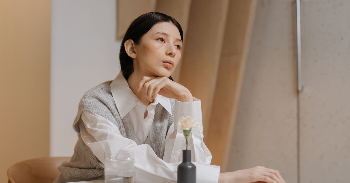 Thoughtful woman in white blouse and vest sitting at a table indoors, lost in deep thought.