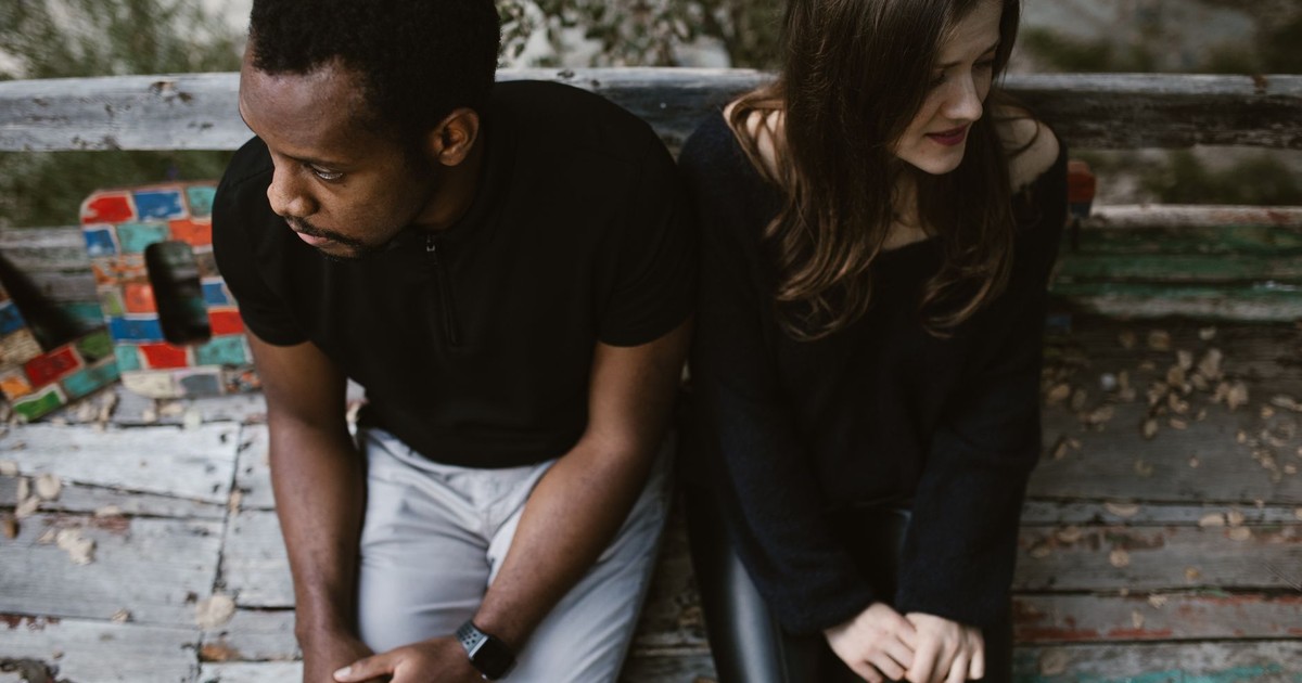 A couple sitting apart on a park bench, expressing emotions. Outdoors setting.