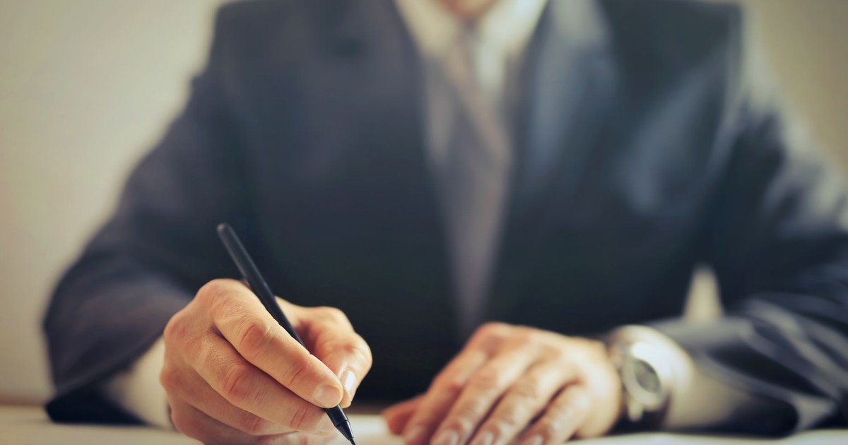 Male entrepreneur wearing formal suit sitting at table in workplace and signing documents while making successful business deal
