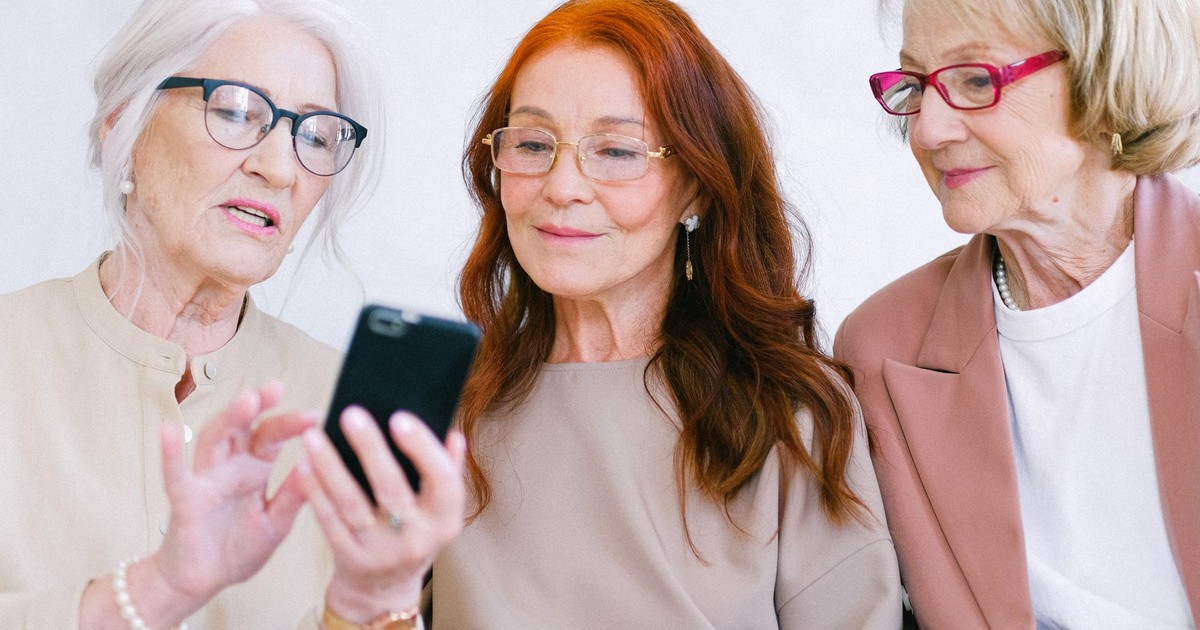 Cheerful senior women talking and using mobile phone while watching video online through internet together