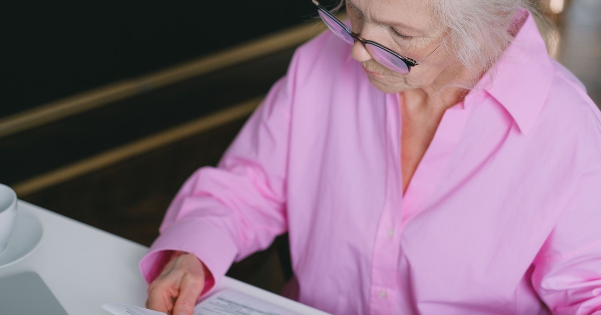 Elderly woman in pink shirt carefully reading a document indoors.