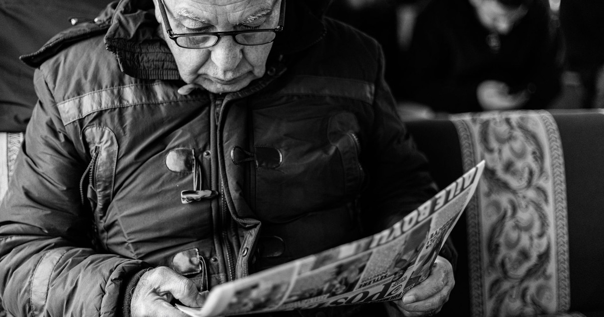 Black and white photo of an elderly man engrossed in newspaper reading on a bus.