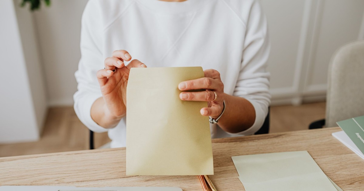 Close-up of a woman sealing a brown envelope while seated at a wooden desk indoors.